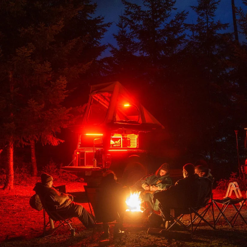 Friends around a campfire at night near a camper lit red by Kingpin lights