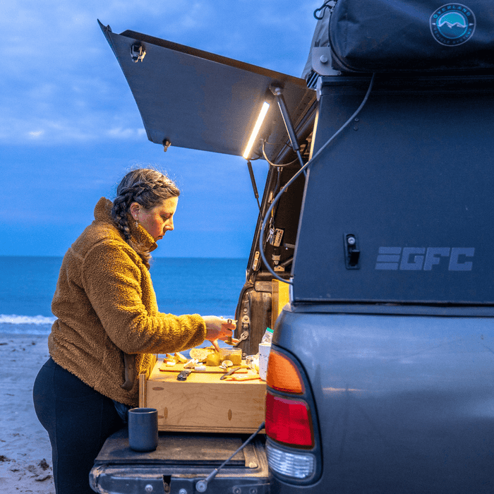 Woman cooking at a truck camper tailgate on the beach under a Kingpin light